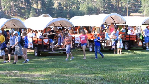 A group of people boarding covered wagons in a grassy area surrounded by trees.