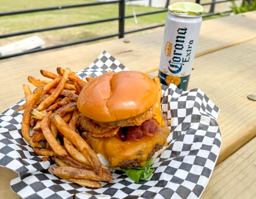 A juicy burger with cheese and bacon, served with crispy fries and a can of Corona on a checkered paper.