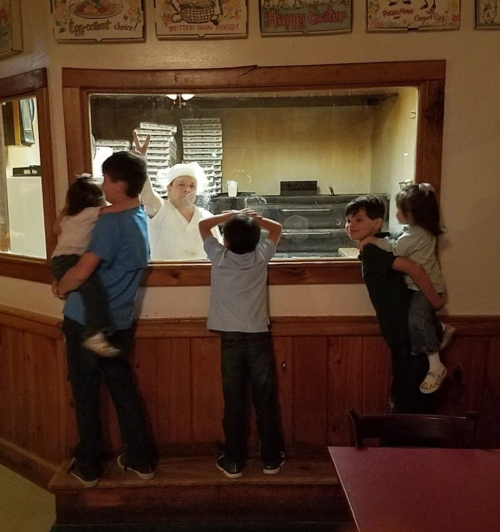 Children watch a chef through a glass window in a restaurant, with some kids being held and others leaning against the wall.