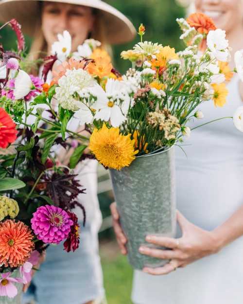 Two people hold vibrant flower bouquets in metal containers, surrounded by greenery on a sunny day.