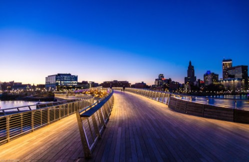 A wooden walkway curves along a river at dusk, with city buildings illuminated in the background.