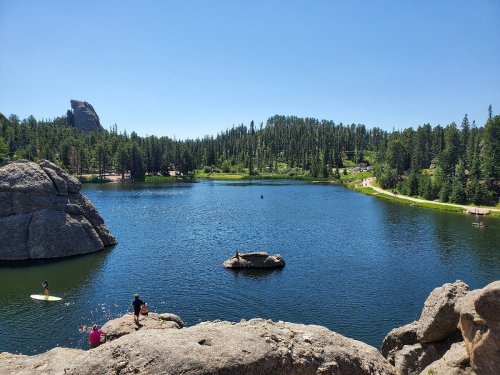 A serene lake surrounded by trees and rocks, with people paddleboarding and enjoying the sunny day.