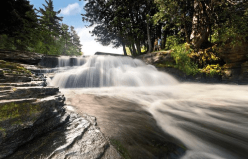 A serene waterfall cascades over rocky ledges, surrounded by lush greenery and trees under a bright sky.