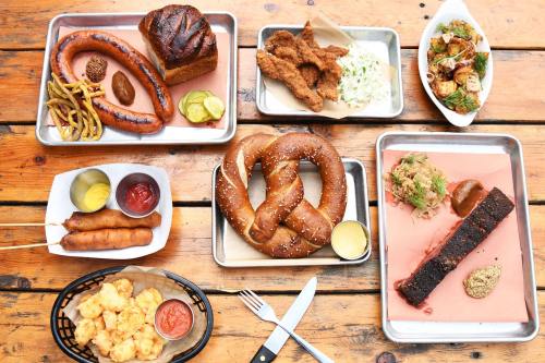 A spread of various dishes including sausages, pretzels, fried chicken, coleslaw, and sides on metal trays.