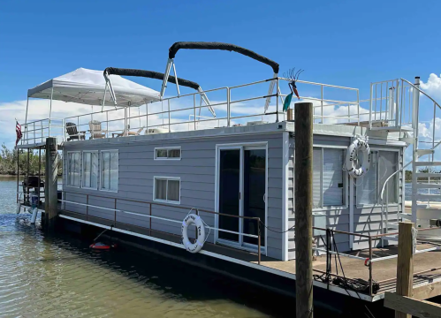 A spacious houseboat with a rooftop deck, moored at a dock on a calm waterway under a clear blue sky.