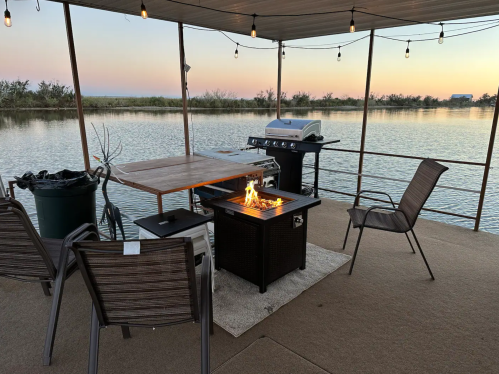 A cozy outdoor space by the water with a fire pit, grill, table, and chairs, illuminated by string lights at sunset.