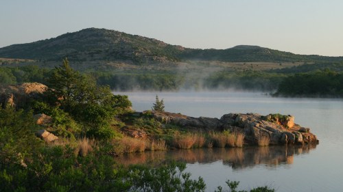 Serene landscape featuring a calm lake, rocky shore, and mist rising in the early morning light with hills in the background.