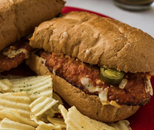 A crispy chicken sandwich with jalapeños on a bun, served with a side of potato chips on a red plate.