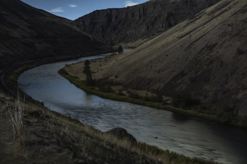 A winding river flows through a rugged canyon, surrounded by steep, rocky hills under a twilight sky.