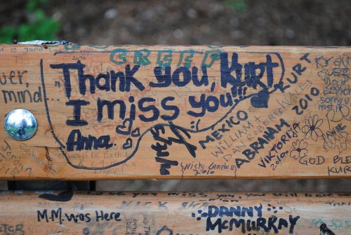A wooden bench covered in graffiti, featuring messages of love and remembrance, including "Thank you, Kurt! I miss you! Ana."