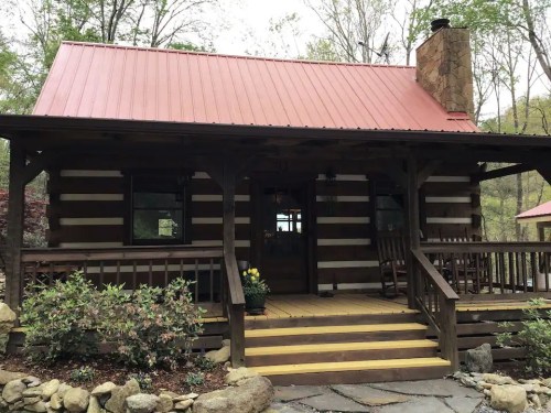 A rustic log cabin with a red metal roof, wooden porch, and stone chimney, surrounded by greenery and flowers.