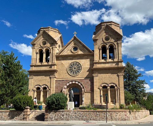 Historic church with twin bell towers, a large clock, and lush greenery under a blue sky with fluffy clouds.