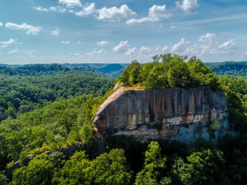 Aerial view of a rocky cliff surrounded by lush green trees and rolling hills under a blue sky with fluffy clouds.