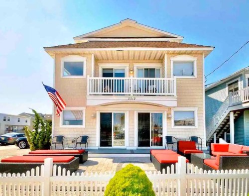 A beach house with a front porch, American flag, and red outdoor seating, surrounded by a white picket fence.