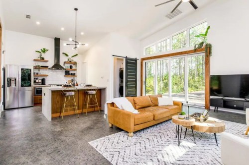 Modern living room with a brown leather sofa, wooden coffee table, and open kitchen featuring plants and large windows.