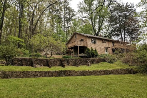 A two-story house surrounded by trees and greenery, with a stone wall and lawn in the foreground.