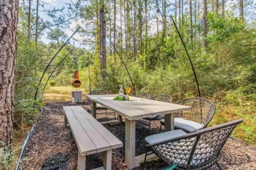 A rustic outdoor dining area with a wooden table, chairs, and a chiminea, surrounded by trees and greenery.
