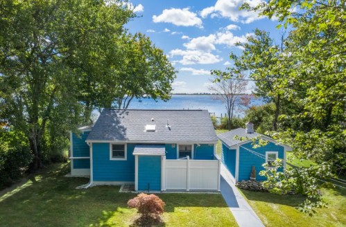A bright blue cottage surrounded by trees, with a view of a lake and a clear blue sky in the background.