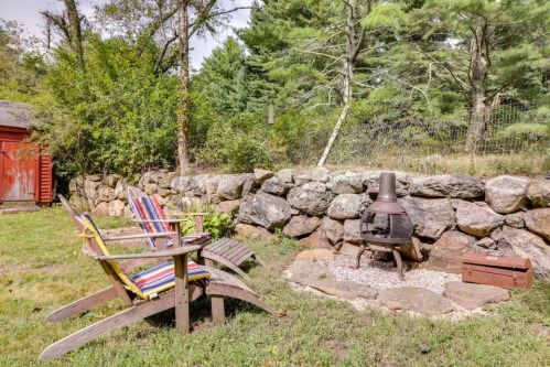 Cozy outdoor seating area with colorful chairs, a fire pit, and a stone wall surrounded by trees.