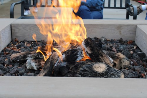 A cozy fire burns in a stone fire pit, surrounded by rocks and wooden logs, with people sitting in the background.