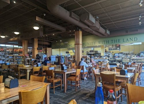 A spacious café with wooden tables and chairs, featuring a community-focused mural and patrons enjoying their time.