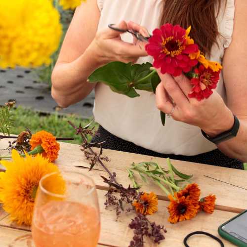 A person arranging colorful flowers on a table, with a drink nearby and various floral materials scattered around.