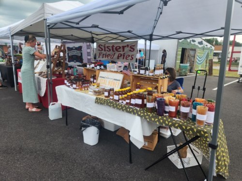 A bustling market scene with stalls selling fried pies and jars of local honey, with shoppers browsing the offerings.