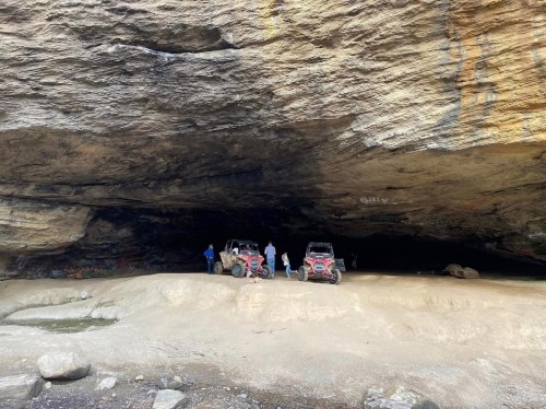 Two off-road vehicles parked under a large rock overhang, with people exploring the area nearby.