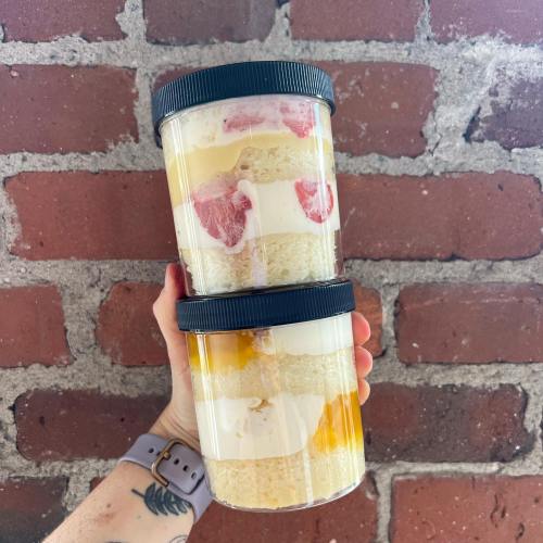 A hand holding two clear containers layered with cake, cream, and fruit against a brick wall background.