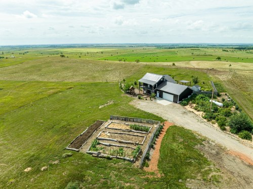 Aerial view of a rural property with a house, garden beds, and expansive green fields under a cloudy sky.