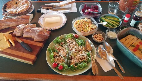 A table spread with various foods: salad, bread, cheese, pie, and colorful vegetables in containers.