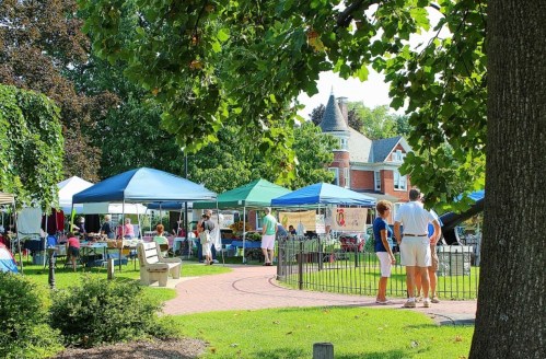 A vibrant outdoor market with colorful tents, people mingling, and greenery in a park setting.