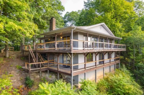 A two-story house with a large deck, surrounded by trees and situated on a hillside.