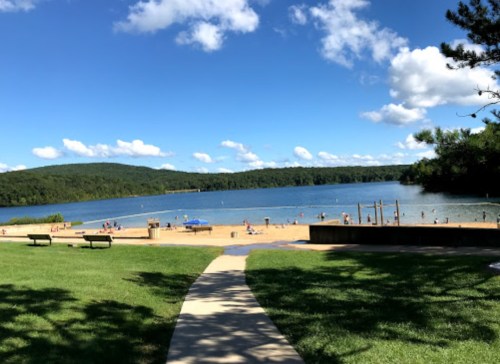 A sunny beach scene with people swimming and relaxing by a lake, surrounded by green hills and blue skies.
