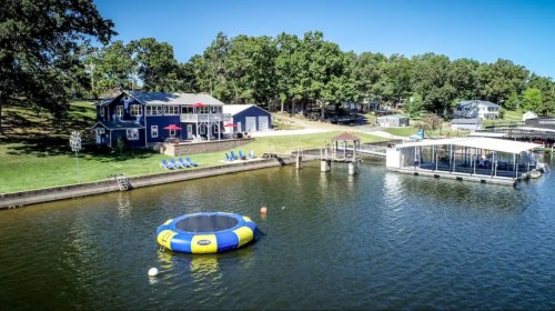 A lakeside view featuring a trampoline, dock, and a blue house surrounded by trees under a clear blue sky.