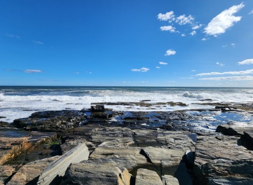 A rocky shoreline with waves crashing against the rocks under a clear blue sky with scattered clouds.