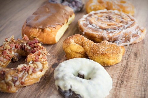 A variety of donuts and pastries arranged on a wooden surface, including glazed, frosted, and bacon-topped options.