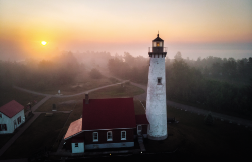 A lighthouse stands tall against a foggy sunrise, with a red-roofed building nearby and trees in the background.