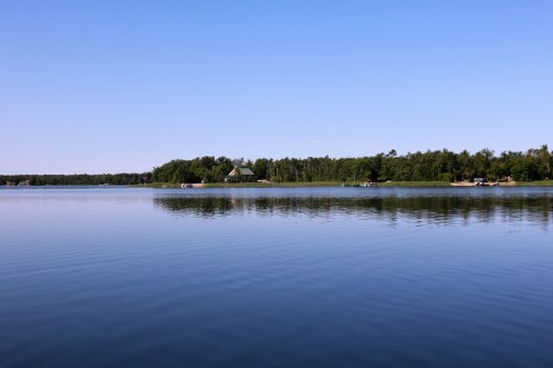 A calm lake reflecting a clear blue sky, surrounded by lush green trees and a distant cabin.
