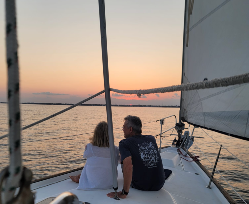 A couple sits on a sailboat, watching a sunset over calm waters, with a serene atmosphere and gentle waves.