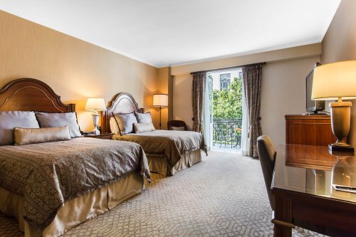 A cozy hotel room with two beds, a desk, and a balcony overlooking greenery. Warm lighting and elegant decor.