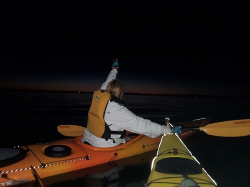A person in a kayak at night raises their hand in a peace sign, with another kayak visible in the foreground.
