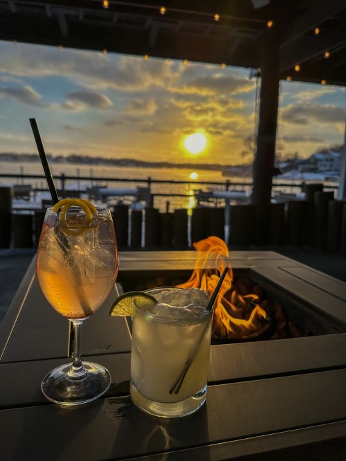 Two cocktails sit on a table by a fire pit, with a sunset over the water in the background.