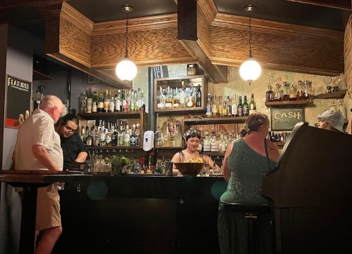 A cozy bar scene with patrons at the counter, a bartender serving drinks, and shelves filled with various bottles.