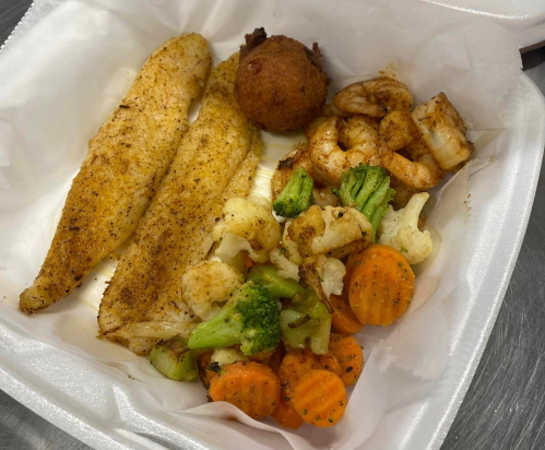 A takeout container with seasoned fish fillets, shrimp, a hushpuppy, and mixed vegetables including carrots and broccoli.