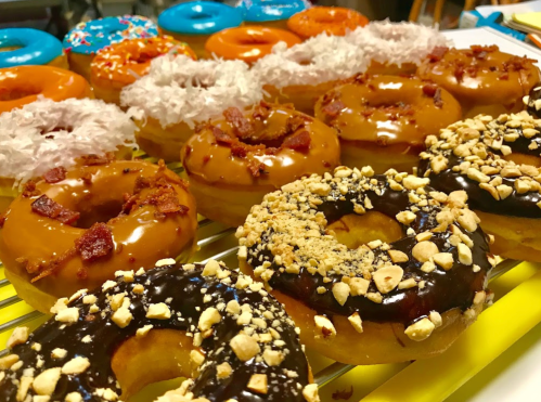 A colorful assortment of donuts, including chocolate, maple bacon, and coconut-topped varieties, arranged on a tray.