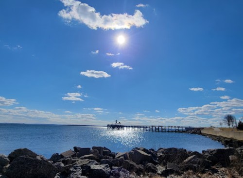 A sunny sky with scattered clouds over a calm sea, featuring a pier and rocky shoreline in the foreground.
