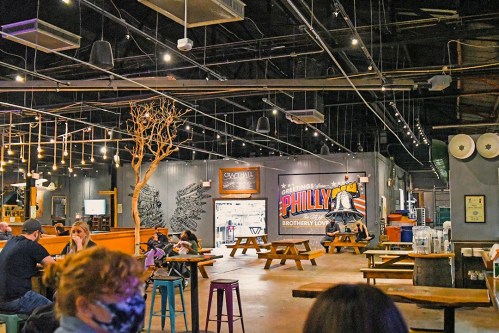 A spacious indoor dining area with wooden tables, string lights, and a mural featuring a Philly cheesesteak theme.
