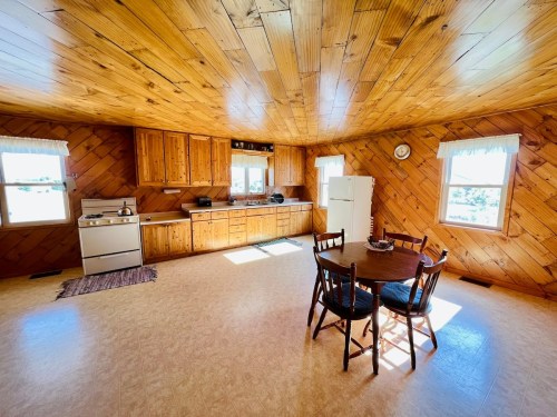 Cozy kitchen with wooden walls, a round table, chairs, and appliances, illuminated by natural light from windows.