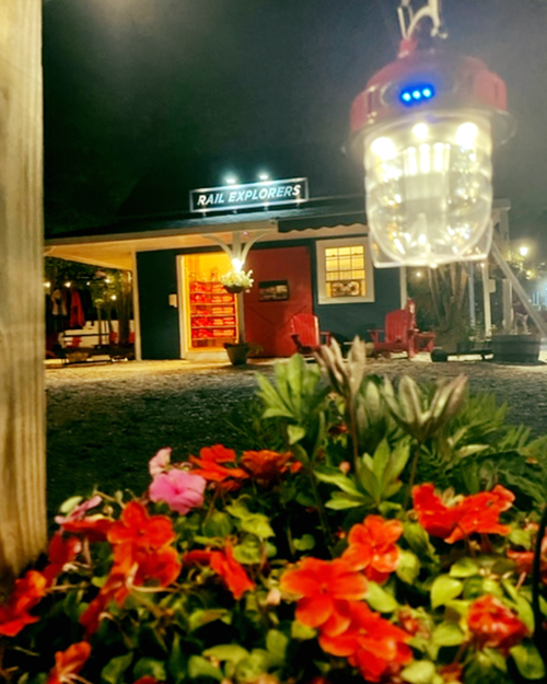 A cozy night scene of a building labeled "Rail Explorers," with colorful flowers in the foreground and a lantern hanging nearby.